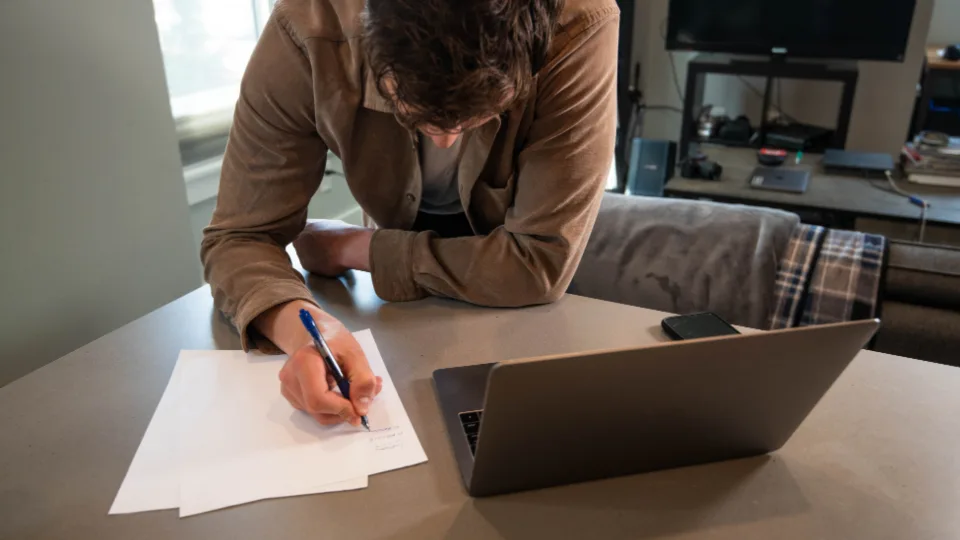 A man leaning over a table, writing notes beside an open laptop