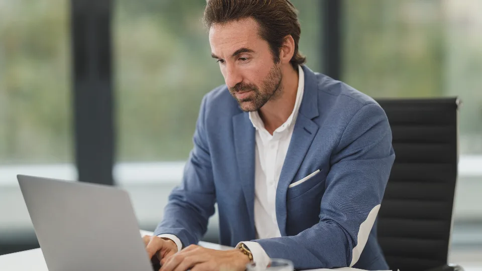 A man in a blazer working at a laptop by an office window
