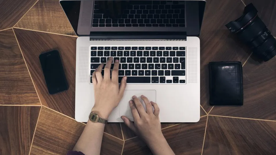 Hands typing on a laptop, seen from above, with a phone and a wallet on the desk